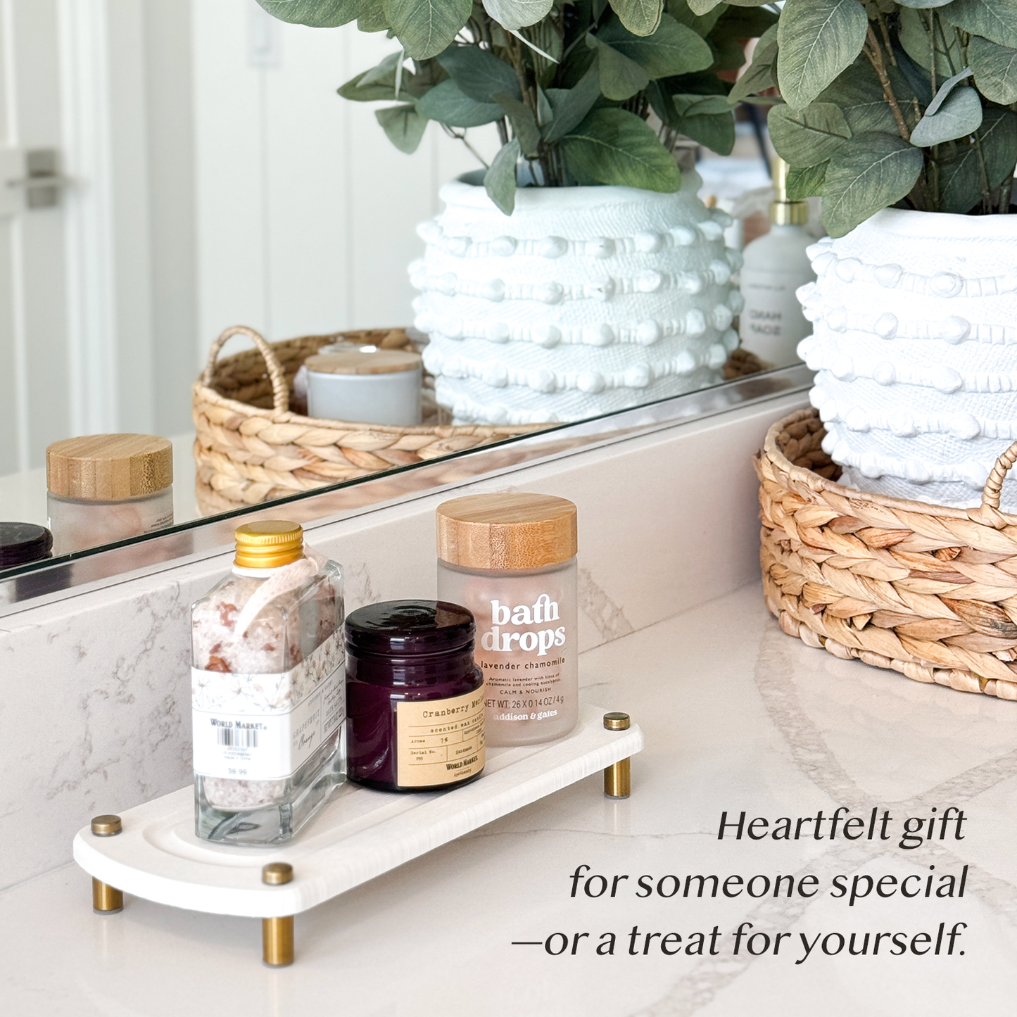 A white sink caddy holding a bath salt jar, a candle, and a bath oil bottle, placed next to bathroom decorations on a countertop.