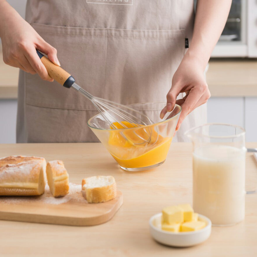 A hand using a balloon whisk to beat eggs in a glass bowl, with a cup of milk, a stick of butter, and a loaf of bread on the countertop nearby.