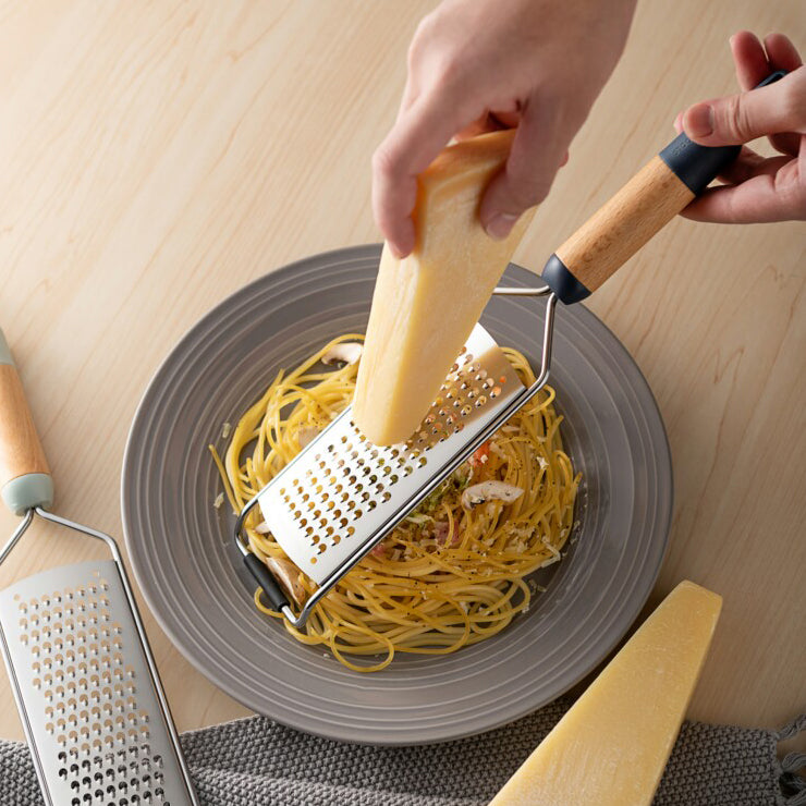 A hand grating cheese over pasta using a midnight blue cheese grater, with freshly grated cheese falling onto the dish.