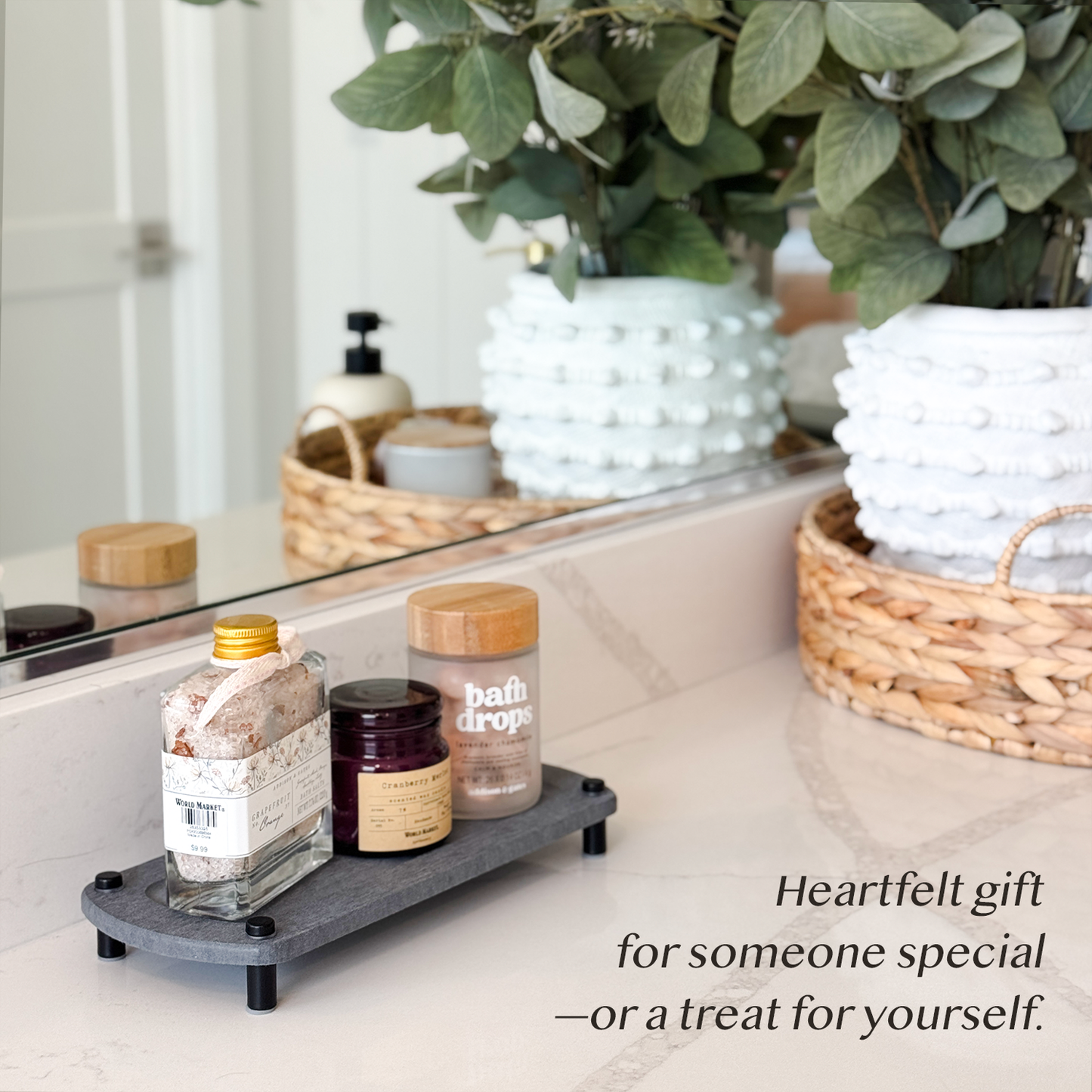 A dark gray sink caddy holding a bath salt jar, a candle, and a bath oil bottle, placed next to bathroom decorations on a countertop.