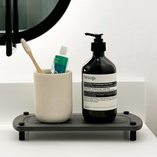 Hand soap and white canister with toothpaste and tooth brush neatly placed on a dark gray sink caddy, set against a bathroom backdrop with a black mirror and white countertop.