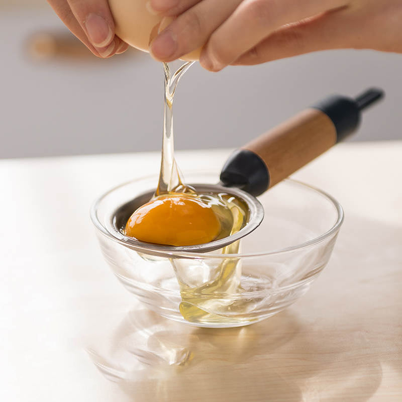 Hands cracking a raw egg over an egg separator; the egg white drips below while the yolk remains in the tool.