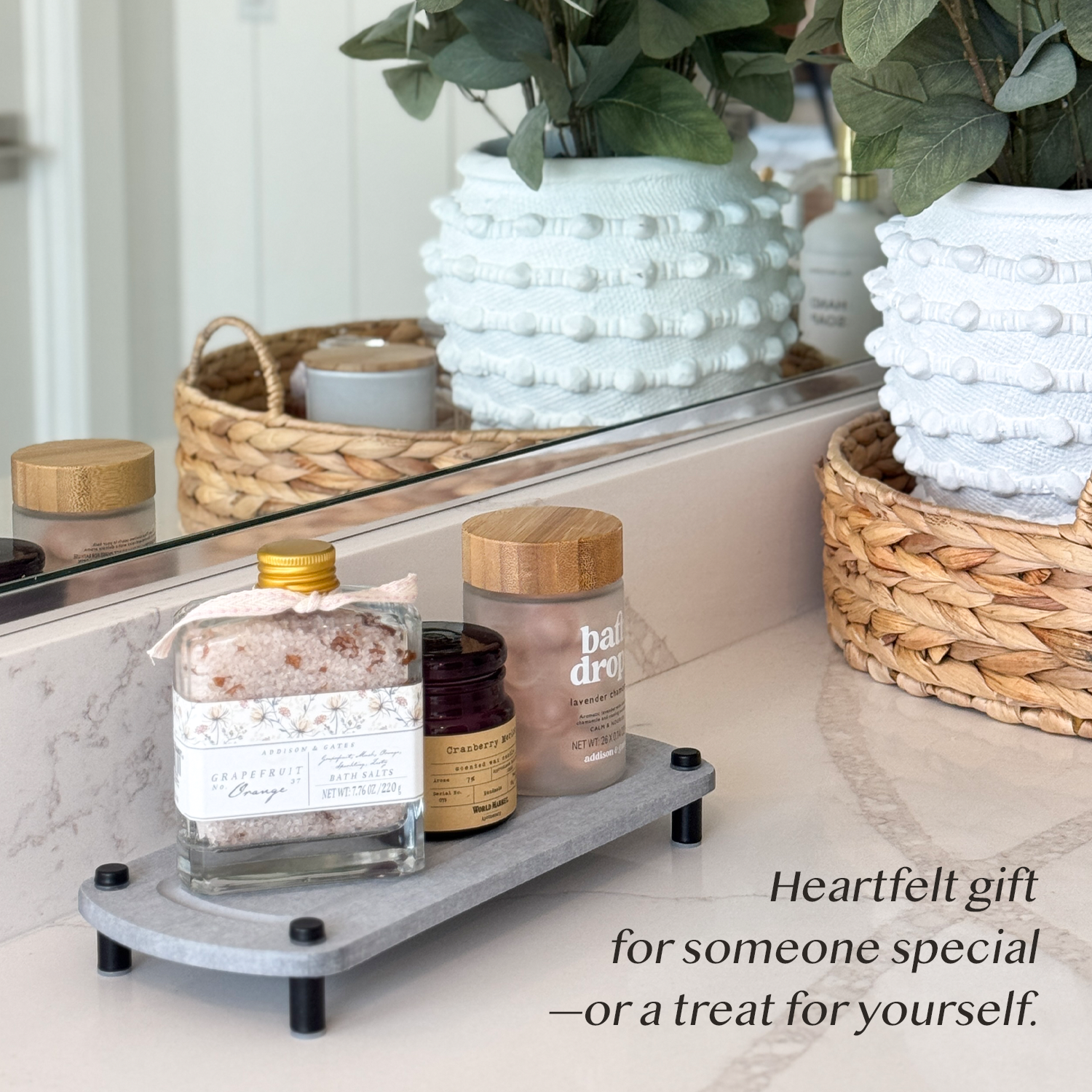 A gray sink caddy holding a bath salt jar, a candle, and a bath oil bottle, placed next to bathroom decorations on a countertop.