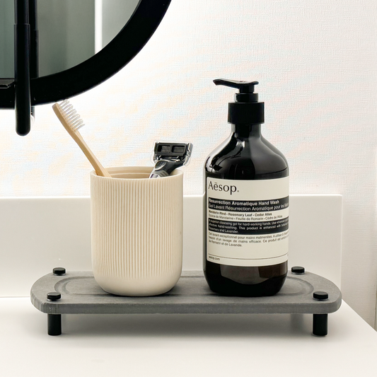 A light gray sink caddy on the bathroom countertop, holding a canister with a toothbrush and razor alongside a hand soap dispenser.