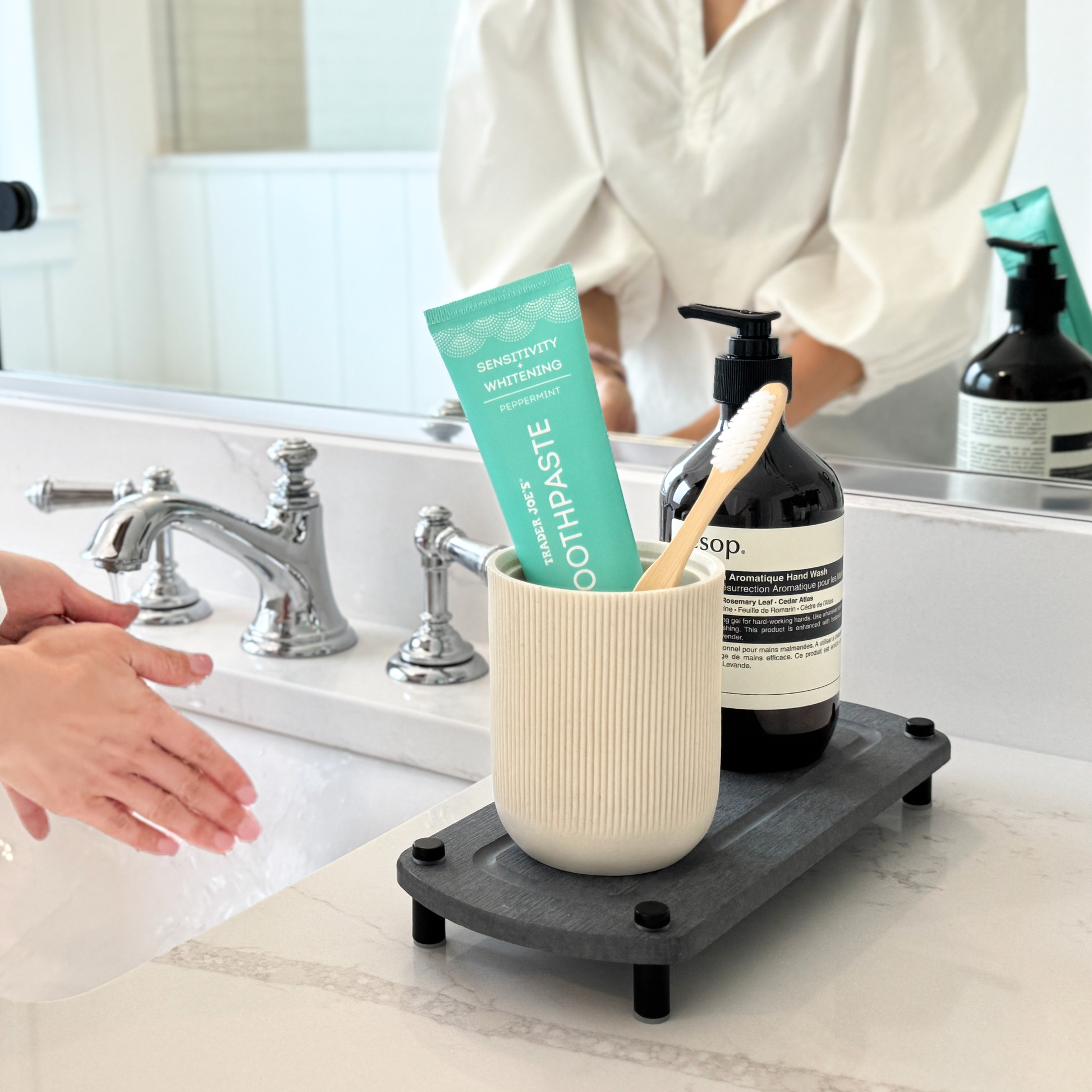 A dark gray sink caddy holding a canister with a toothbrush and toothpaste, along with a hand soap dispenser, placed next to a woman washing her hands at the sink.