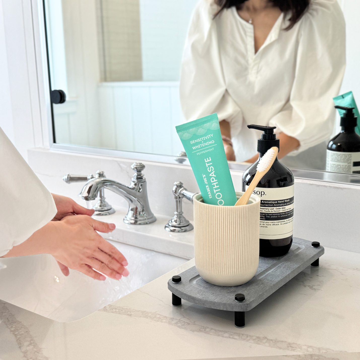 A glacier gray sink caddy holding a canister with a toothbrush and toothpaste, along with a hand soap dispenser, placed next to a woman washing her hands at the sink.