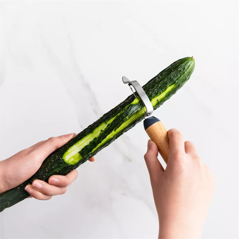 A hand smoothly peeling a long cucumber using an I-shaped peeler, with thin strips of green skin curling away, revealing the pale flesh beneath.