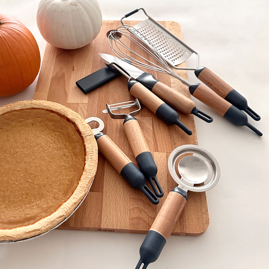 A 7-piece kitchen tool set, including a paring knife, Y-shaped peeler, I-shaped peeler, balloon whisk, cheese grater, egg separator, and bottle opener, is neatly arranged on a wooden chopping board. Adjacent to the board, a freshly baked pumpkin pie and small decorative pumpkins evoke a cozy autumn ambiance.
