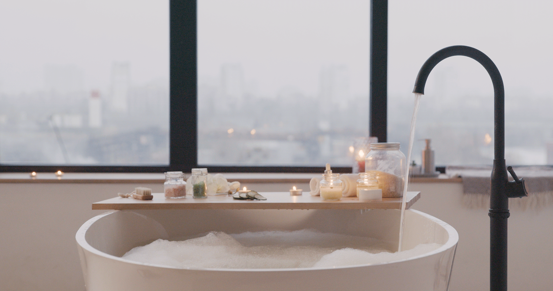 Cozy bathroom scene with water flowing from a sleek black faucet, candles flickering, and a serene view of a steam-filled stream through a large window.