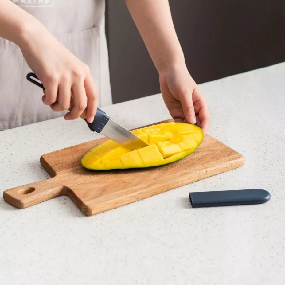 A hand uses a paring knife to slice a ripe mango in half on a wooden chopping board, with the knife's protective case placed nearby.