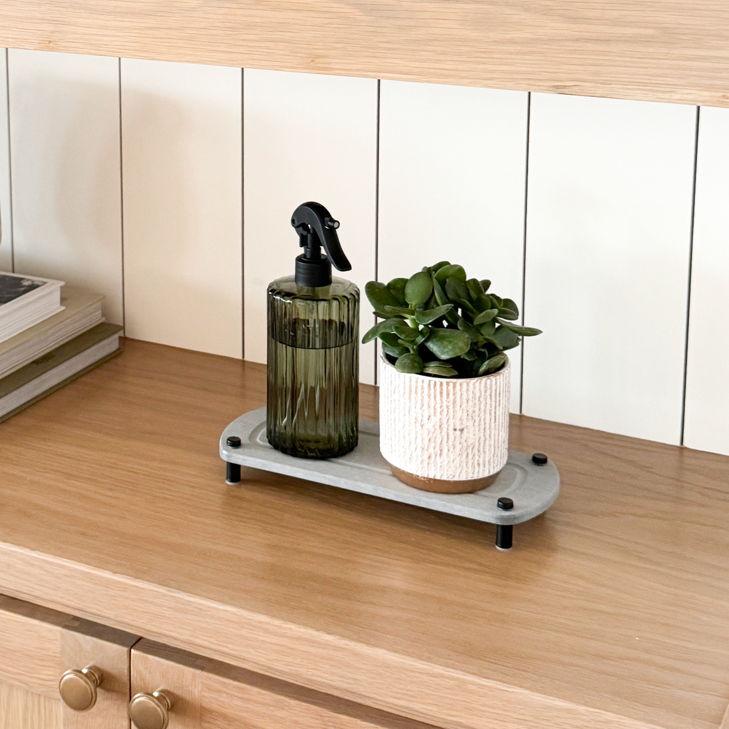 A gray sink caddy on a console, with a water spray bottle and a pot placed on it, surrounded by a candle and various plants.
