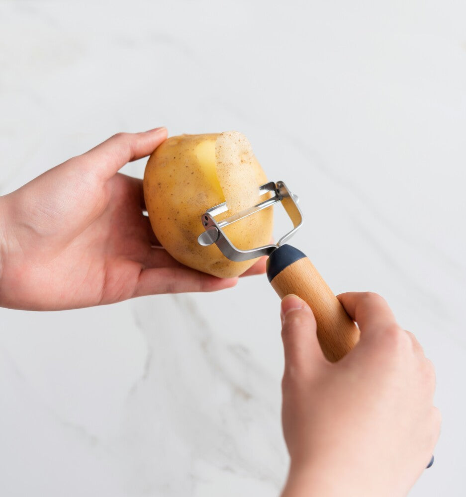 A hand smoothly peeling a potato with a Y-shaped peeler, removing thin strips of skin to reveal the clean surface beneath.